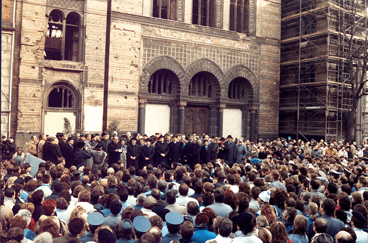 Foto einer Gruppe von Menschen in dunklen, langen Mänteln und größtenteils mit dunklen Hüten steht erhöht vor einer großen Menschenmenge auf der Freifläche im Hof der zerstörten Synagoge in Mitte. Eine Person auf dem Podest (Mitte-links) hält eine Rede. Hinter der Gruppe ist das historische Gebäude zu sehen, die Fenster fehlen, einige Türen sind zubetoniert, die Fassade sieht deutlich zerstört aus, der rechte Teil ist von einem Baugerüst umgeben.