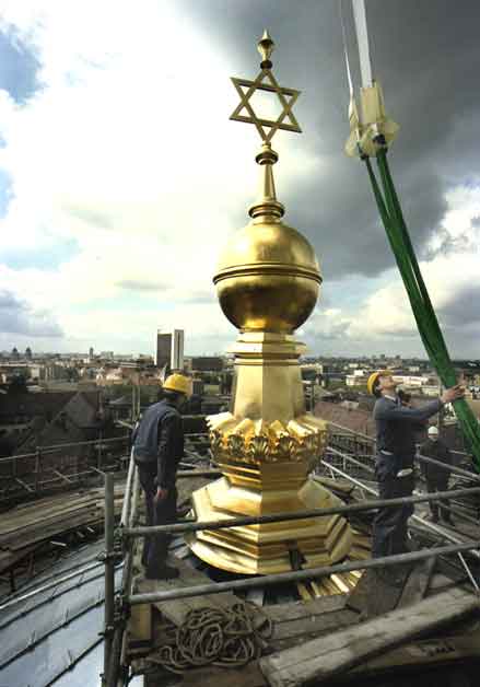 Foto von der goldenen Spitze, welche die Kuppel der Neuen Synagoge schmückt. Das Foto wurde aus einer leichten Draufsicht auf die Kuppel aufgenommen. Zwei Personen in blauen Anzügen und gelben Schuzthelmen stehen auf einem Gerüst, das den Fuß der Spitze umzäunt. Die Person rechts im Bild greift mit der rechten Hand um grüne Seile, die an einem Kran befestigt sind, der allerdings nicht im Bild zu sehen ist. Im Hintergrund sind Häuserdächer zu erkennen.