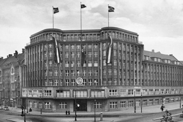 Schwarz-weiß Fotografie vom Kaufhaus Jonass, es handelt sich um ein Eckgebäude mit acht Etagen. Das Foto zeigt eine Draufsicht von der gegenüberliegenden Straßenseite auf die Vorderseite des Gebäudes und einen Ausschnitt der Straßenkreuzung vor dem Gebäude. Auf dem Dach und auf Höhe der obersten Etage sind Fahnen der Nationalsozialistischen Partei gehisst.