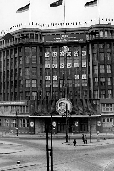 Hochformatige schwarz-weiß-Fotografie des Zentralhauses der Einheit. Das Foto zeigt eine Draufsicht auf das Eckgebäude, oben auf dem Dach sind zwei Deutschlandfahnen und eine sowjetische Fahne gehisst. Auf Höhe der obersten Etage ist ein Banner angebracht mit der Aufschrift "XXXIV - Es lebe der 24. Jahrestag der grossen sozialistischen Oktoberrevolution".