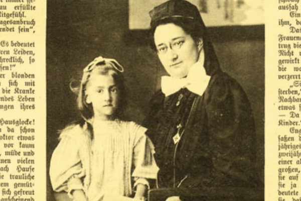 Gelbliche schwarz-weiß Fotografie von Henriette Arendt (rechts) mit Kind (links), die in einer Zeitung abgedruckt ist. Henriette Arendt trägt ein dunkles, langärmliges Gewand und eine weiße Schleife um den Hals, eine runde Brille sowie eine dunkle Kopfbedeckung. Das Kind trägt ein helles Kleid mit Puffärmeln und eine helle Schleife auf dem Kopf. Beide sitzen und schauen in die Kamera. Der Bildausschnitt zeigt nut die Oberköper der Personen.