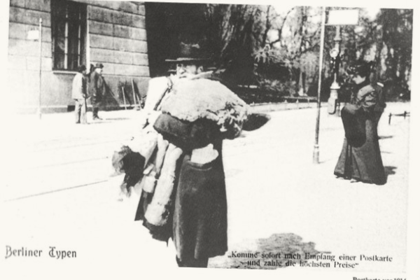 Schwarz-weißes Foto eines jüdischen Altkleiderhändlers in Berlin, der mit einem großen Sack auf der Schulter geht. Im Hintergrund sind Passanten und Gebäude sichtbar.